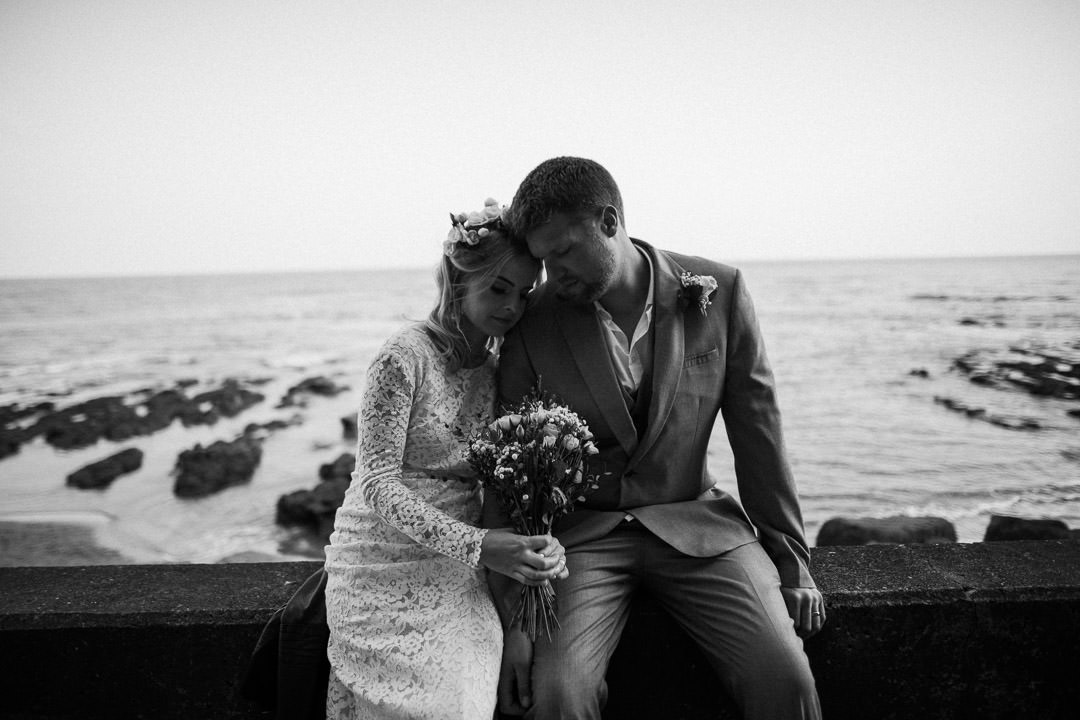 bride and groom sat on wall near ocean
