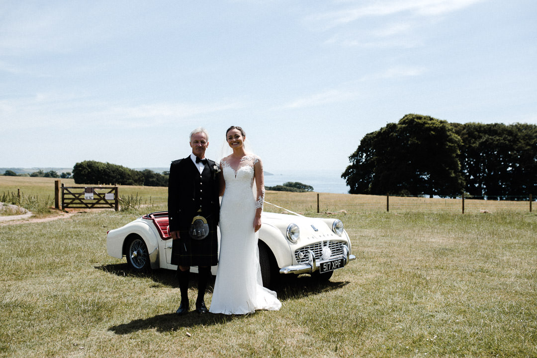 bride and her father stood next to classic car in a field