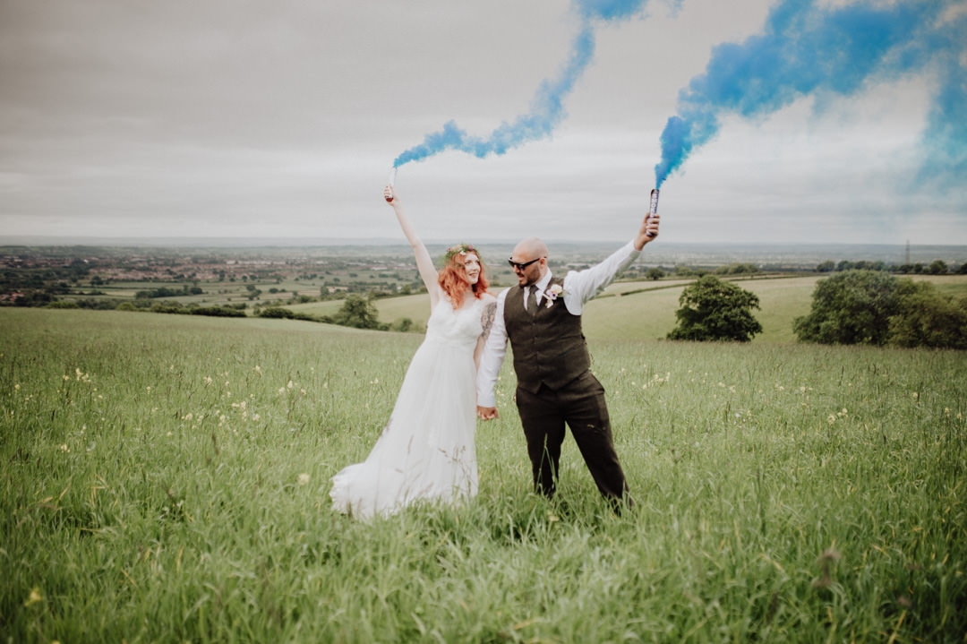 bride and groom holding smoke flares in meadow