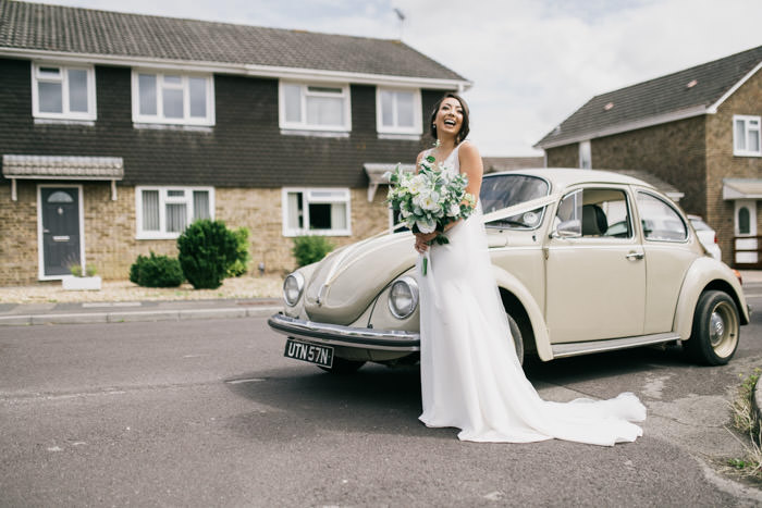 bride standing in a dress next to classic car holding wedding flowers in street