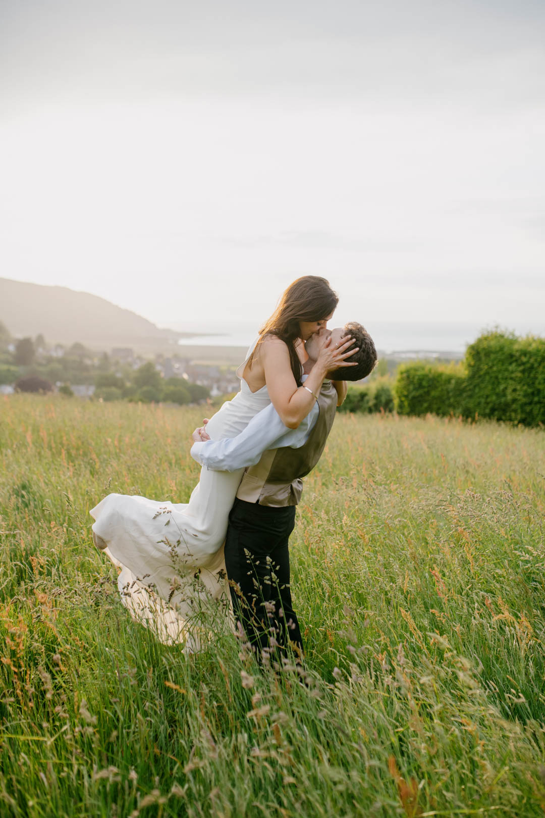 groom lifting up bride in meadow kissing her during day time