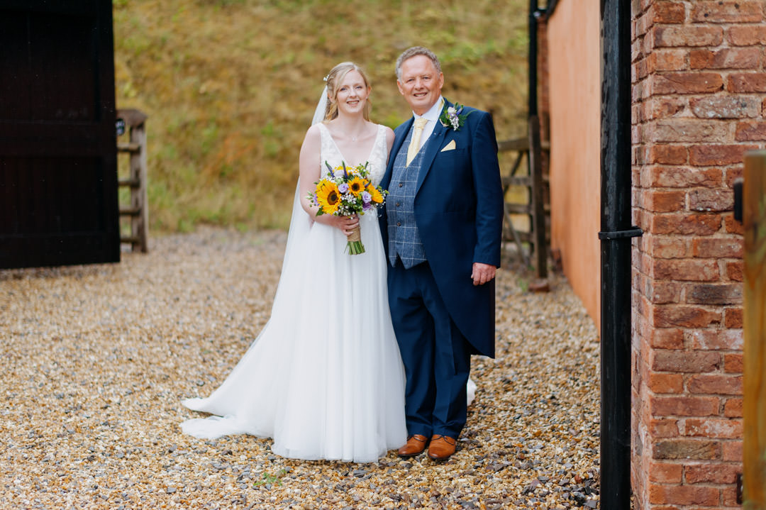 father and bride stood next to large barn holding sunflowers