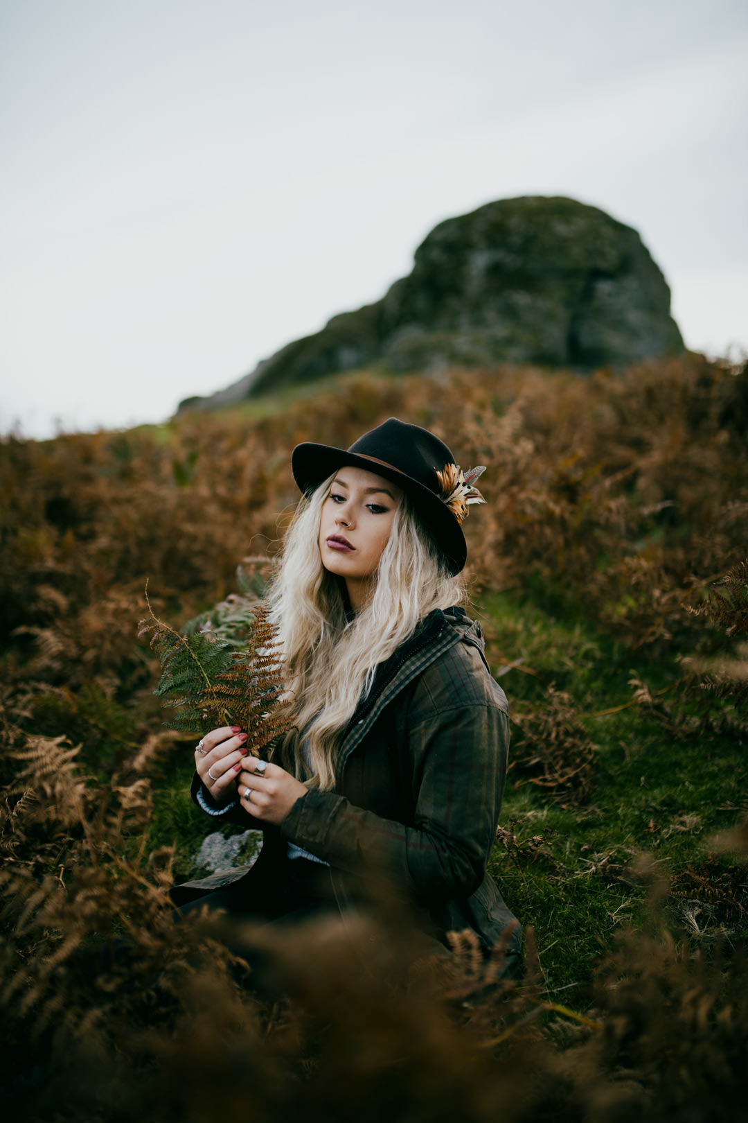 blonde hair woman waring hat sat in long grass near mountain