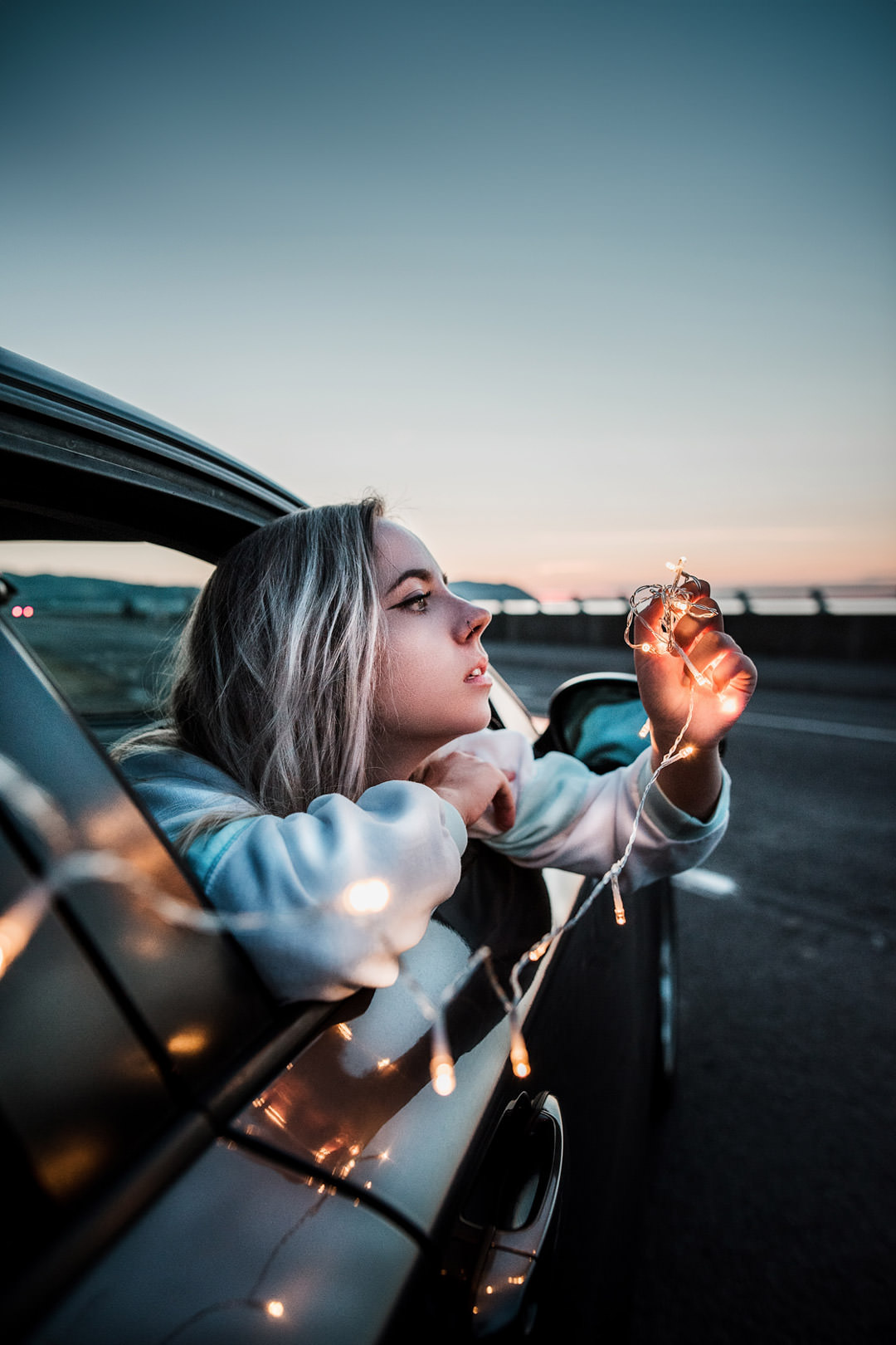girl with blonde hair leaning out car window holding string lights