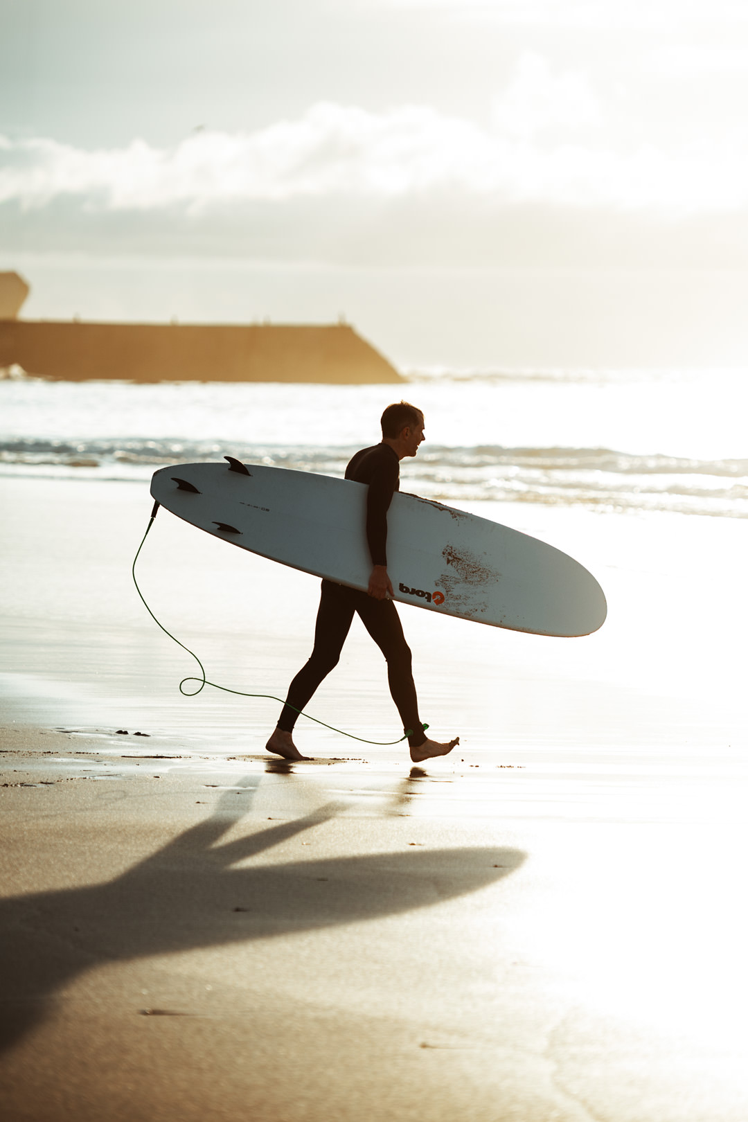 man carrying surfboard towards ocean on flat sandy beach during sunset