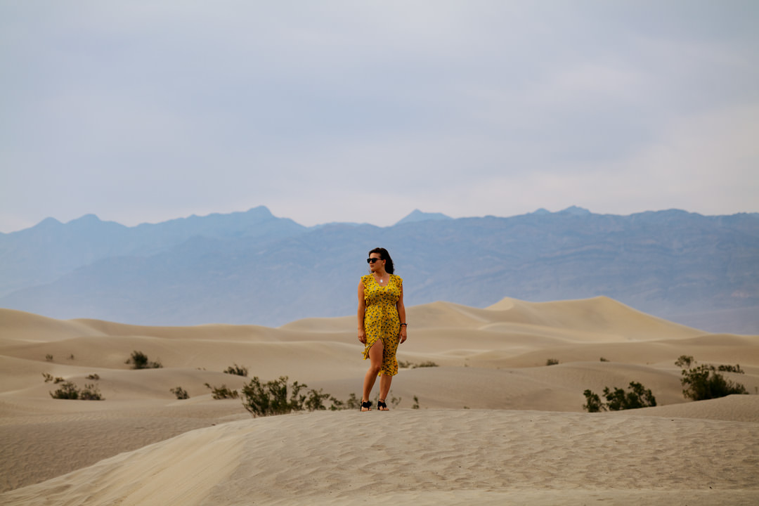 girl in yellow dress stood on large sand dune under blue sky