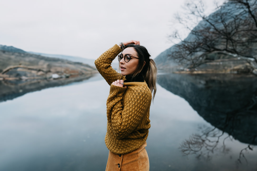 girl waring orange jumper stood next to large frozen lake near mountains