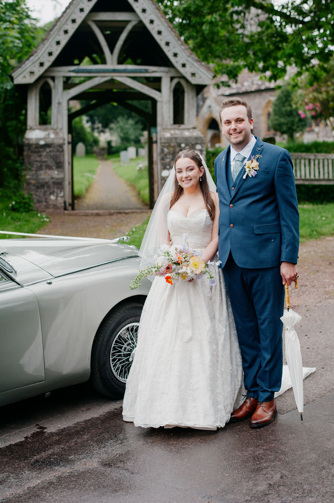 bride and groom stood in road next to grey classic sports car