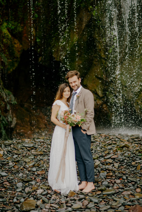 bride and groom stood next to large waterfall on beach