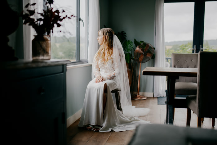bride sat on stool in window light waring white wedding dress
