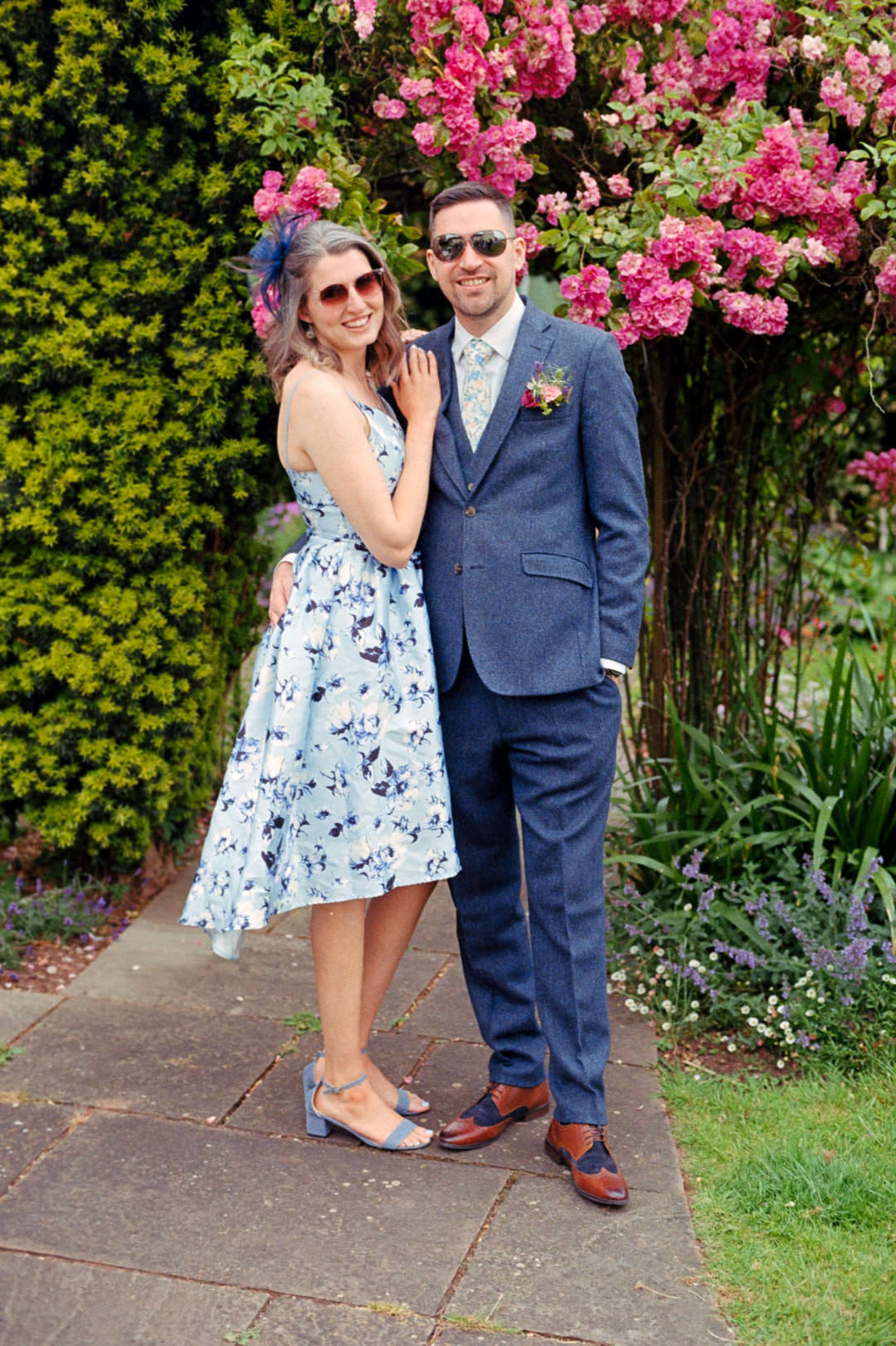 young couple holding hands stood under large rose bush during day time
